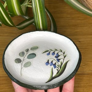 Könnte beinhalten: Eine kleine, ovale Keramikschale, weiß mit einem dunkelgrünen Rand. Die Schale ist mit bemalten botanischen Designs verziert, darunter ein Zweig mit blauen Blumen und eine Gruppe grüner Blätter. Die Schale wird in einer Hand gehalten.