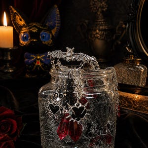 May include: A clear glass jar filled with silver-toned jewelry, including charms shaped like bats and hearts with black enamel. Several pieces feature dangling, faceted red crystal pendants. A silver bat-shaped ornament rests on the jar's rim.