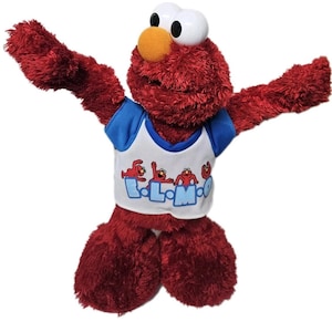 May include: A red Elmo soft toy with large white eyes, an orange nose, and a blue and white shirt. The shirt features the text "Elmo" and several Elmo figures. The toy's arms are outstretched.