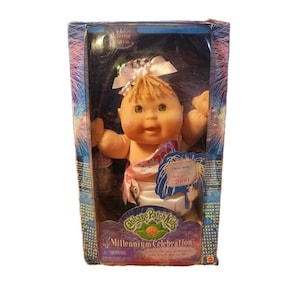 Puede incluir: Muñeca Cabbage Patch Kids Millennium Celebration en su embalaje original. La muñeca tiene cabello rubio, un lazo blanco y una faja rosa. El embalaje presenta fuegos artificiales y el texto "Millennium Celebration".