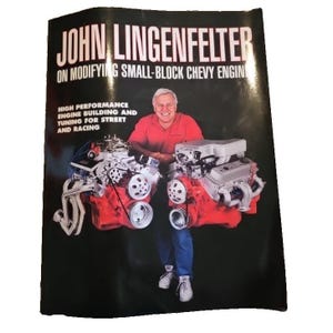 May include: A book cover with the title "JOHN LINGENFELTER" and the subtitle "ON MODIFYING SMALL-BLOCK CHEVY ENGIN". The cover also includes the text "HIGH PERFORMANCE ENGINE BUILDING AND TUNING FOR STREET AND RACING". The image shows a man in a red shirt and blue jeans with two car engines.