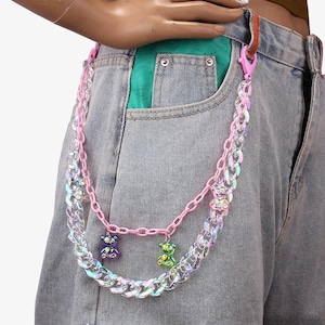 May include: A pink and iridescent chain with two small gummy bear charms. The chain is attached to a pair of light wash denim jeans with a green pocket lining. The chain is a fashion accessory.