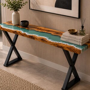 May include: A long console table featuring a natural wood slab top with a central turquoise resin river inlay that mimics a shoreline. The table is supported by two black metal X-shaped legs. The surface holds a white vase, a small bowl, and books.