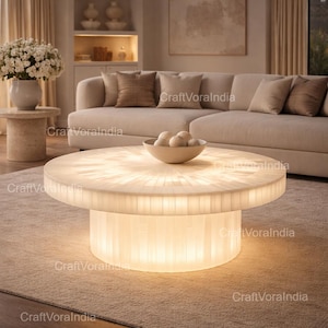 May include: A circular, illuminated coffee table with a white, mosaic-style top and a cylindrical base. The table is the focal point of a living room setting, with a neutral colour palette and soft lighting. A bowl with decorative spheres sits on the table.