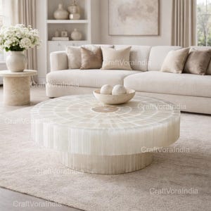 Puede incluir: Una mesa de centro redonda y blanca con una parte superior de mosaico y una base cilíndrica. La mesa está en un salón con un sofá blanco, cojines beige y un jarrón con flores blancas. La mesa es un punto focal.