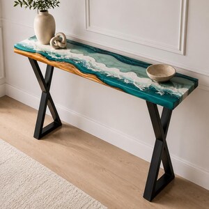 May include: A console table featuring a live edge wood slab top with a teal and white resin inlay mimicking ocean waves. The table is supported by two black metal X-shaped legs. A beige vase with greenery and a small bowl sit on the surface.