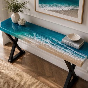 Puede incluir: Una mesa consola larga con una base de metal negro en forma de X y una superficie rectangular con un diseño de resina que imita una costa oceánica con agua azul, olas de espuma blanca y una textura de playa de arena.