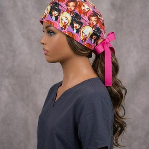 May include: A scrub cap featuring a repeating pattern of stylised doll faces with large eyes and vibrant hair colours on a pink background. The cap is secured at the back with a bright pink ribbon tied into a bow, providing a snug fit for the wearer.