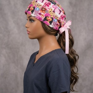 May include: A pink scrub cap featuring a repeating Bratz doll graphic print with various makeup and fashion icons. The cap includes a light pink ribbon tie at the back to secure the fit, showcasing a vibrant collage of pink, yellow, and black.