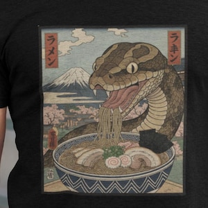 Könnte beinhalten: Schwarzes T-Shirt mit einem japanisch inspirierten Motiv einer Schlange, die Ramen isst. Das Design umfasst eine Schüssel Nudeln, eine Berglandschaft und japanischen Text. Das T-Shirt ist aus einem weichen Material.