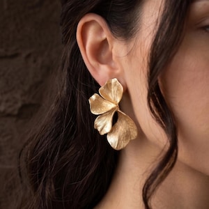 May include: Gold-coloured floral earrings. The earrings feature multiple textured petals, forming a three-dimensional flower shape. The earrings are worn on the ear, with the back of the ear and part of the face visible.