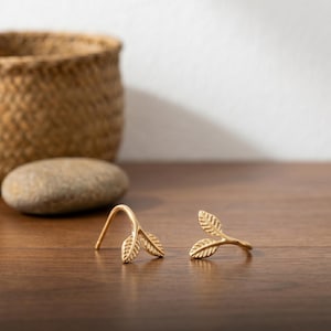 Op de afbeelding: Een paar goudkleurige oorbellen in de vorm van kleine, delicate takjes met elk twee gestructureerde blaadjes. De oorbellen hebben een gebogen stiftontwerp, wat zorgt voor een elegante uitstraling met een gepolijste metalen afwerking en fijne bladnerven.