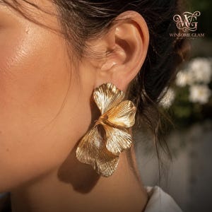May include: A gold-toned earring featuring a layered, fan-like design with a textured, ribbed surface that mimics the appearance of organic petals or leaves. The piece has a metallic finish and is secured to the earlobe with a post backing.