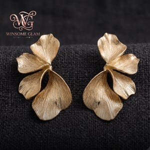 Può includere: Un paio di orecchini color oro caratterizzati da un design floreale testurizzato a più petali con sottili striature verticali. Gli orecchini hanno una finitura metallica spazzolata e una forma organica asimmetrica. Il logo WINSOME GLAM è visibile.