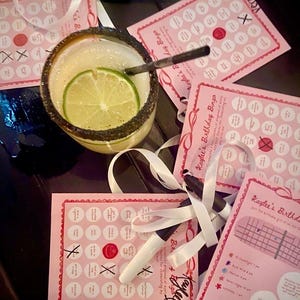 Puede incluir: Un cóctel de margarita con una rodaja de lima y un vaso con borde de sal, junto a tarjetas de bingo rosas de cumpleaños con círculos blancos y detalles rojos. También hay rotuladores negros y cintas blancas.
