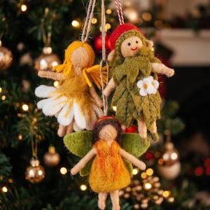 May include: Three needle-felted fairy ornaments hanging by twine. One has yellow hair and a white petal skirt, one wears a red cap and green leaf outfit with a white flower, and one has brown hair, green wings, and a yellow dress.