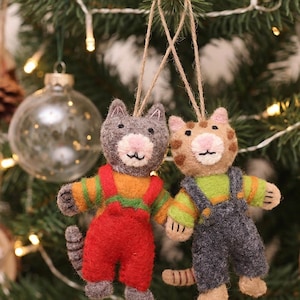 May include: Two needle-felted cat ornaments hanging from jute twine. The gray cat wears red overalls over an orange and green striped shirt. The tan tabby cat wears dark gray overalls over a lime green and orange striped shirt.