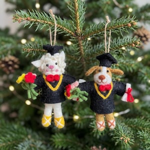 May include: Two handmade graduation ornaments. One is a white sheep, the other a brown and white dog. Both wear black graduation gowns with yellow trim and red bow ties. Each holds a bouquet of flowers and a diploma. They hang from a Christmas tree.