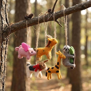 May include: A collection of felt animal ornaments, including a pig with pink wings, two giraffes, a dog with a red scarf, and a sloth holding a leaf. The ornaments are suspended from a tree branch with twine.
