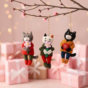 Peut inclure: Trois ornements de chat faits à la main suspendus à une branche avec des fleurs roses. Les chats sont vêtus de tenues colorées et tiennent des fleurs ou un cœur. L'arrière-plan comprend des boîtes cadeaux roses.