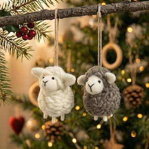 May include: Two hanging ornaments shaped like sheep, one white and one dark grey, crafted from textured yarn with felted faces, black bead eyes, and stitched noses, each suspended by a tan twine loop from a horizontal wooden branch.