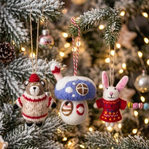 May include: Three felt ornaments hanging from a tree: a brown bear wearing a beige and red knit sweater, a blue mushroom house with a white snowy top, and a white rabbit in a red dress holding a string of colorful beads on a thin cord.