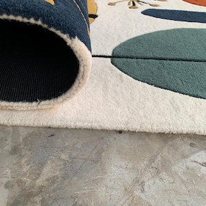 Puede incluir: Una alfombra enrollada con un dise&ntilde;o moderno de mediados de siglo. La alfombra presenta un fondo crema con formas abstractas en verde azulado, naranja, amarillo y azul marino. El borde de la alfombra revela un reverso azul marino.