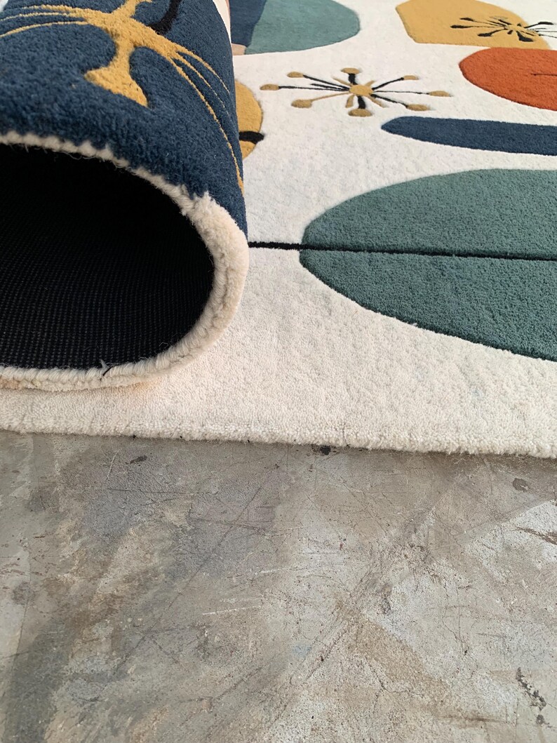 Puede incluir: Una alfombra enrollada con un dise&ntilde;o moderno de mediados de siglo. La alfombra presenta un fondo crema con formas abstractas en verde azulado, naranja, amarillo y azul marino. El borde de la alfombra revela un reverso azul marino.