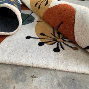 Puede incluir: Una alfombra enrollada con un dise&ntilde;o moderno de mediados de siglo. La alfombra presenta un fondo crema con formas geom&eacute;tricas en naranja, amarillo, verde azulado y azul marino. Los detalles florales negros a&ntilde;aden detalles al dise&ntilde;o.