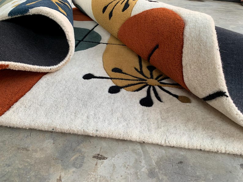Puede incluir: Una alfombra enrollada con un dise&ntilde;o moderno de mediados de siglo. La alfombra presenta un fondo crema con formas geom&eacute;tricas en naranja, amarillo, verde azulado y azul marino. Los detalles florales negros a&ntilde;aden detalles al dise&ntilde;o.