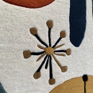 Puede incluir: Primer plano de una alfombra moderna de mediados de siglo con un fondo crema. La alfombra presenta formas abstractas en &oacute;xido, azul marino y amarillo mostaza. Un dise&ntilde;o de estrella en marr&oacute;n y negro a&ntilde;ade un toque &uacute;nico al dise&ntilde;o de la alfombra.