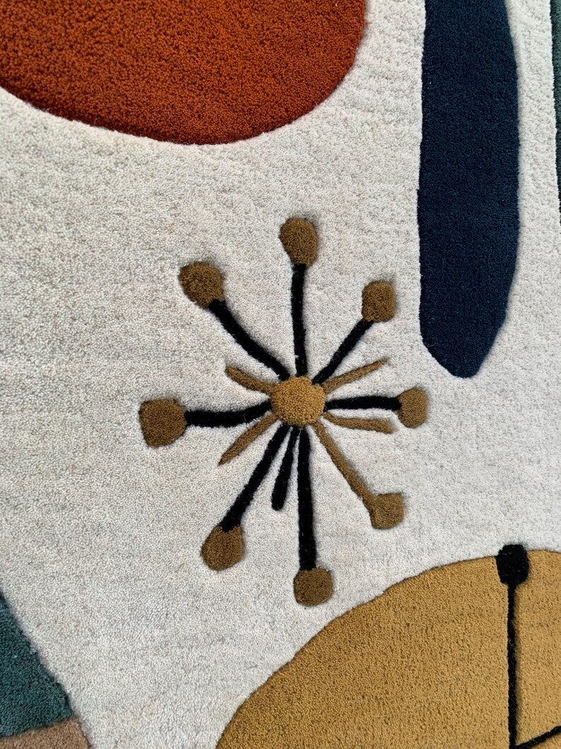Puede incluir: Primer plano de una alfombra moderna de mediados de siglo con un fondo crema. La alfombra presenta formas abstractas en &oacute;xido, azul marino y amarillo mostaza. Un dise&ntilde;o de estrella en marr&oacute;n y negro a&ntilde;ade un toque &uacute;nico al dise&ntilde;o de la alfombra.