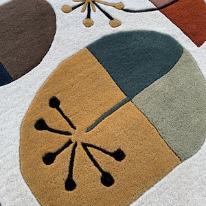 Puede incluir: Una alfombra abstracta con fondo blanco y formas geom&eacute;tricas en marr&oacute;n, naranja, rosa y verde. La alfombra presenta un &oacute;valo grande segmentado y dise&ntilde;os de estrella en negro y dorado.