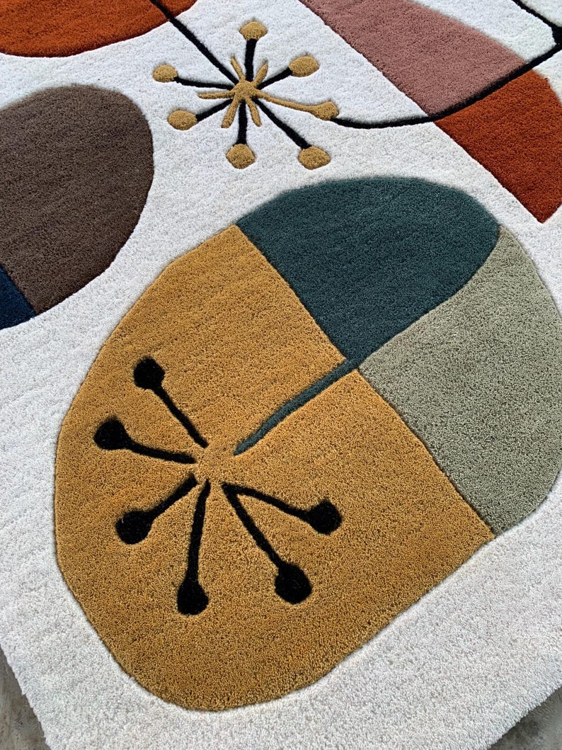 Puede incluir: Una alfombra abstracta con fondo blanco y formas geom&eacute;tricas en marr&oacute;n, naranja, rosa y verde. La alfombra presenta un &oacute;valo grande segmentado y dise&ntilde;os de estrella en negro y dorado.
