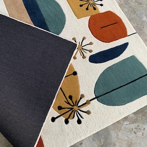 Puede incluir: Una alfombra rectangular con fondo crema y formas abstractas coloridas. El dise&ntilde;o incluye formas en verde azulado, azul marino, naranja y amarillo mostaza, con detalles lineales negros. La parte inferior de la alfombra es azul marino oscuro.
