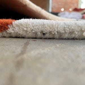 Puede incluir: Primer plano de una secci&oacute;n de una alfombra con una pila texturizada y en bucle. La alfombra presenta un dise&ntilde;o de bloques de color con secciones blancas y naranjas. La alfombra est&aacute; sobre una superficie gris claro.