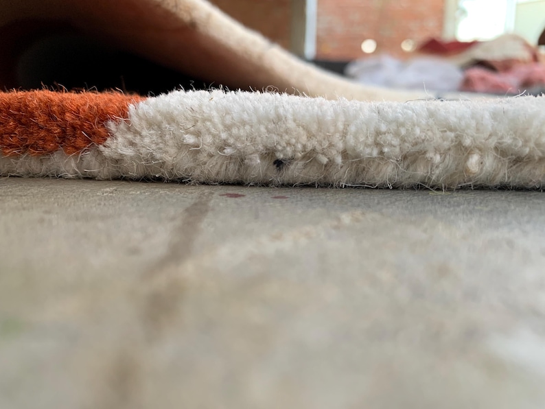 Puede incluir: Primer plano de una secci&oacute;n de una alfombra con una pila texturizada y en bucle. La alfombra presenta un dise&ntilde;o de bloques de color con secciones blancas y naranjas. La alfombra est&aacute; sobre una superficie gris claro.