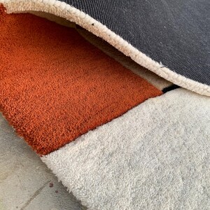 Puede incluir: Primer plano de una alfombra tupida con un dise&ntilde;o geom&eacute;trico. La alfombra presenta una secci&oacute;n naranja, blanca y negra. El reverso de la alfombra es gris oscuro. La alfombra tiene un aspecto suave y texturizado.