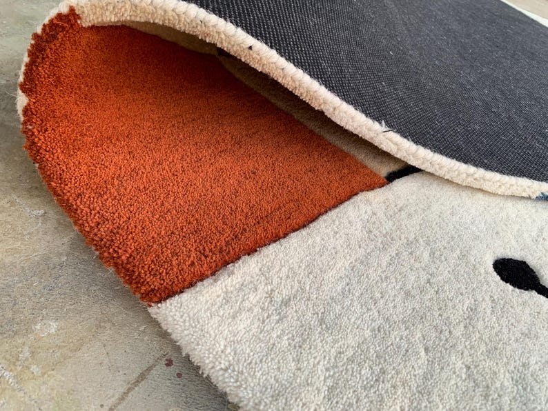 Puede incluir: Primer plano de una alfombra tupida con un dise&ntilde;o geom&eacute;trico. La alfombra presenta una secci&oacute;n naranja, blanca y negra. El reverso de la alfombra es gris oscuro. La alfombra tiene un aspecto suave y texturizado.