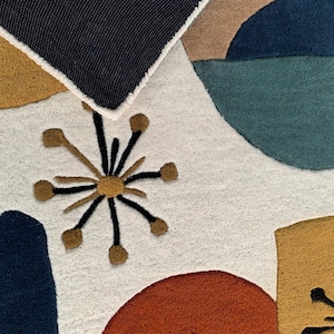 Puede incluir: Primer plano de una colorida alfombra abstracta con fondo blanco. La alfombra presenta formas geom&eacute;tricas en tonos de azul, verde azulado, naranja y dorado. Una secci&oacute;n del borde azul oscuro de la alfombra es visible en la parte superior.