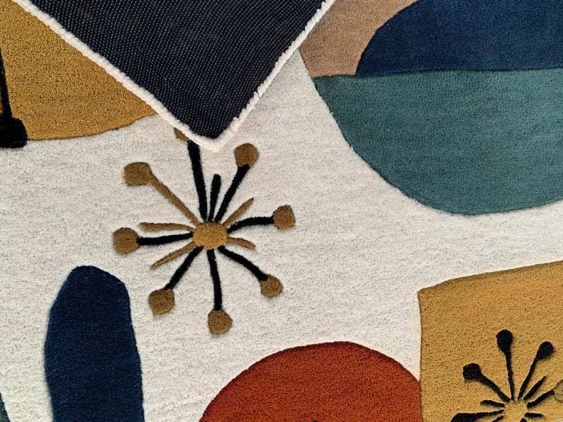 Puede incluir: Primer plano de una colorida alfombra abstracta con fondo blanco. La alfombra presenta formas geom&eacute;tricas en tonos de azul, verde azulado, naranja y dorado. Una secci&oacute;n del borde azul oscuro de la alfombra es visible en la parte superior.