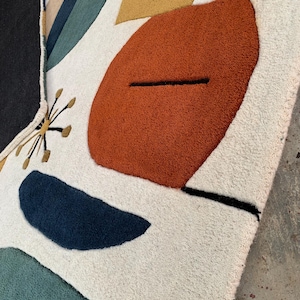 Puede incluir: Una alfombra color crema con un dise&ntilde;o abstracto. La alfombra presenta grandes formas texturizadas en &oacute;xido, azul marino, verde azulado y amarillo mostaza, con detalles en negro y dorado. El dise&ntilde;o es moderno y geom&eacute;trico, con una textura suave y afelpada.