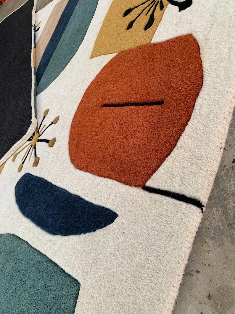 Puede incluir: Una alfombra color crema con un dise&ntilde;o abstracto. La alfombra presenta grandes formas texturizadas en &oacute;xido, azul marino, verde azulado y amarillo mostaza, con detalles en negro y dorado. El dise&ntilde;o es moderno y geom&eacute;trico, con una textura suave y afelpada.