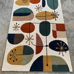 Puede incluir: Una alfombra color crema con un dise&ntilde;o moderno de mediados de siglo. La alfombra presenta varias formas en tonos de naranja, amarillo, verde y azul, conectadas por l&iacute;neas negras y detalles de estrellas. La alfombra es rectangular y parece estar hecha de un material suave.