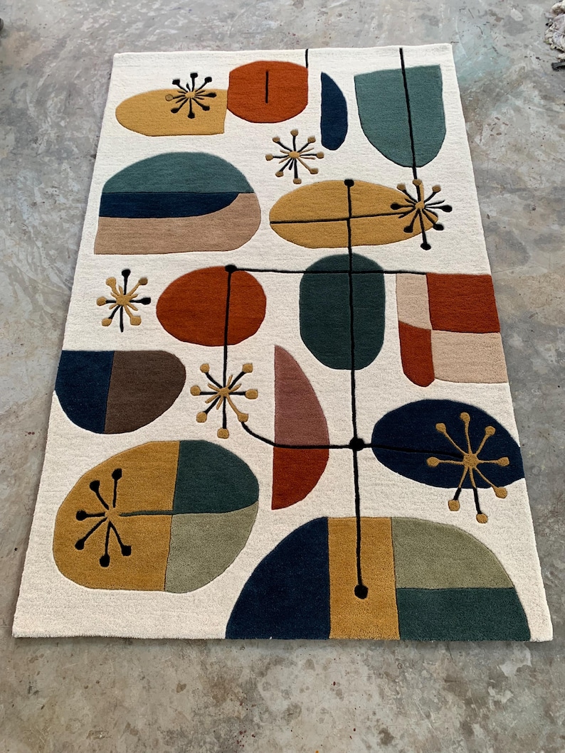Puede incluir: Una alfombra color crema con un dise&ntilde;o moderno de mediados de siglo. La alfombra presenta varias formas en tonos de naranja, amarillo, verde y azul, conectadas por l&iacute;neas negras y detalles de estrellas. La alfombra es rectangular y parece estar hecha de un material suave.