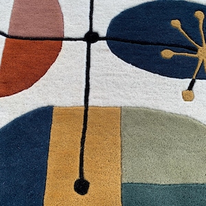 Puede incluir: Primer plano de una colorida alfombra con un dise&ntilde;o abstracto. La alfombra presenta formas en tonos de azul, naranja, rosa y dorado sobre un fondo blanco. Una l&iacute;nea negra con forma circular intersecta el dise&ntilde;o, a&ntilde;adiendo un toque moderno.