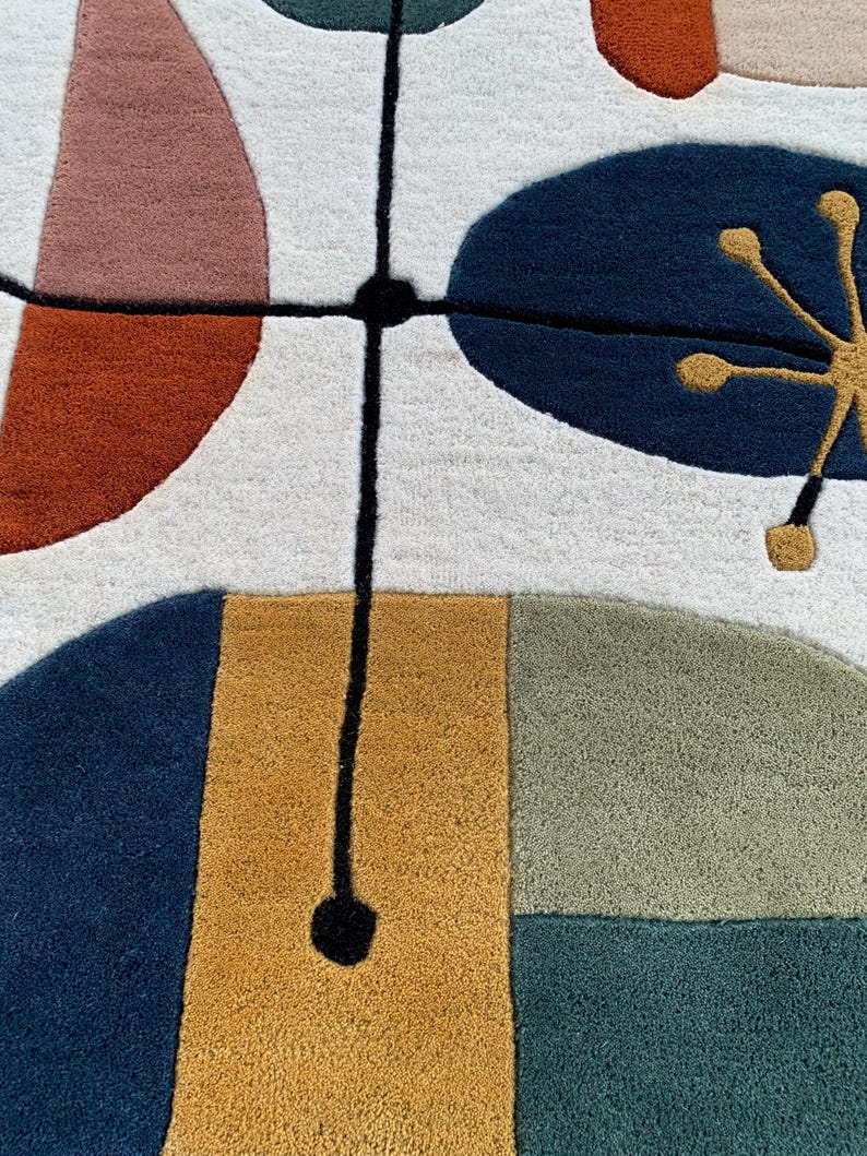 Puede incluir: Primer plano de una colorida alfombra con un dise&ntilde;o abstracto. La alfombra presenta formas en tonos de azul, naranja, rosa y dorado sobre un fondo blanco. Una l&iacute;nea negra con forma circular intersecta el dise&ntilde;o, a&ntilde;adiendo un toque moderno.
