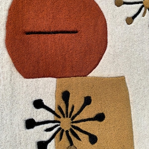 Puede incluir: Primer plano de una alfombra color crema con un dise&ntilde;o moderno de mediados de siglo. La alfombra presenta formas grandes y texturizadas en &oacute;xido, dorado y negro. El dise&ntilde;o incluye un &oacute;valo color &oacute;xido con una l&iacute;nea negra y formas de estrella.