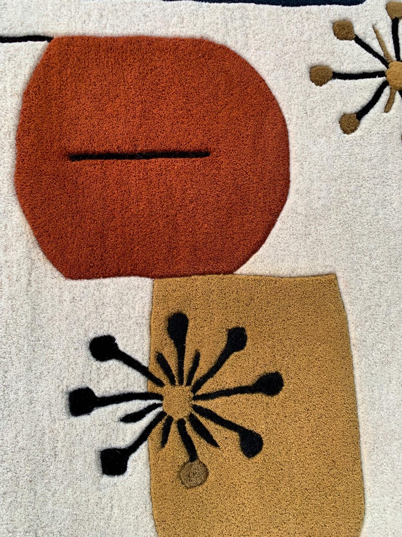 Puede incluir: Primer plano de una alfombra color crema con un dise&ntilde;o moderno de mediados de siglo. La alfombra presenta formas grandes y texturizadas en &oacute;xido, dorado y negro. El dise&ntilde;o incluye un &oacute;valo color &oacute;xido con una l&iacute;nea negra y formas de estrella.