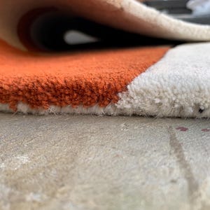 Puede incluir: Primer plano de una alfombra redonda enrollada con una superficie texturizada. La alfombra presenta un dise&ntilde;o de bloques de color con secciones naranjas y blancas. El borde de la alfombra es visible, mostrando el grosor y la textura del material.
